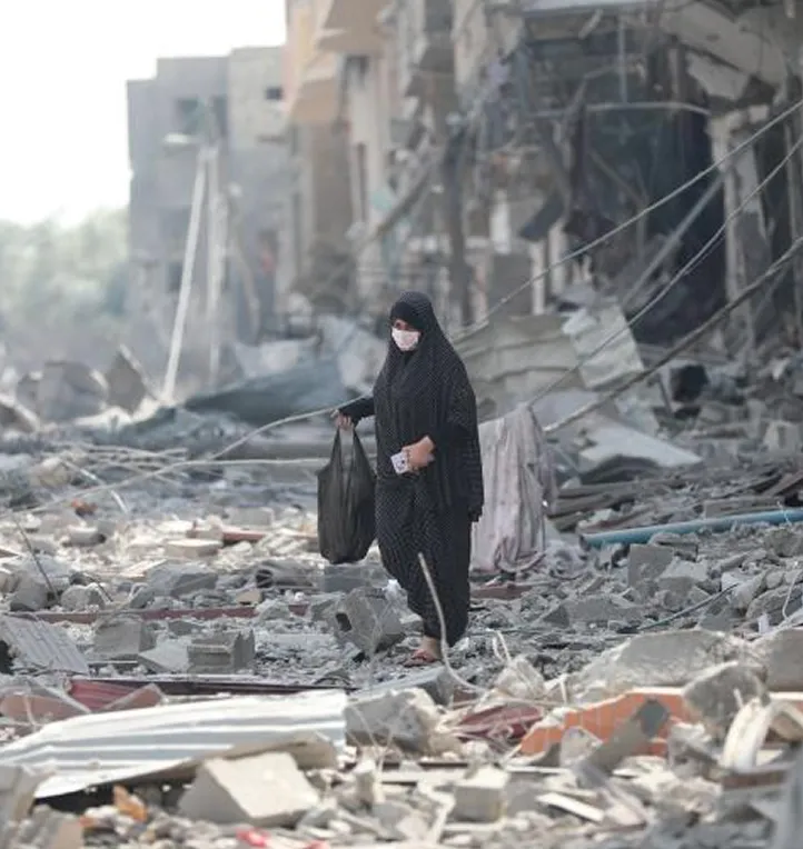 Woman walking through rubble of destroyed buildings, wearing a mask and carrying a bag.