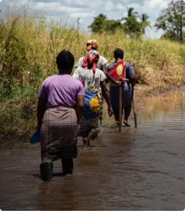 Women walking through a flooded rural path, carrying goods, under a cloudy sky.