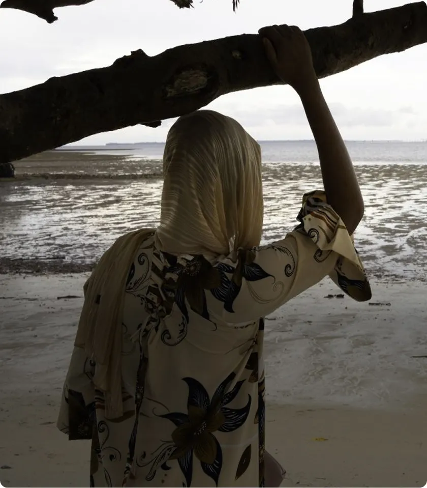 Person in floral attire and headscarf gazing at serene beach landscape under a tree branch.