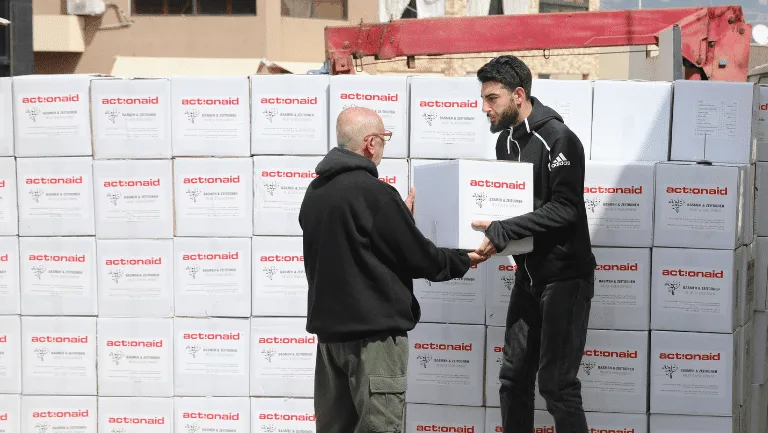 Two people distribute ActionAid relief boxes from a stack during a humanitarian aid effort.