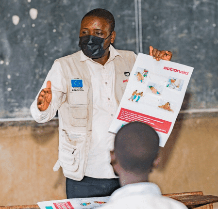 Instructor discusses educational material in classroom setting, wearing a mask, showing booklet to students.