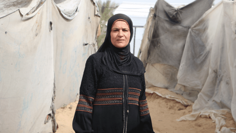 Woman in traditional attire stands in front of canvas tents at a refugee camp.