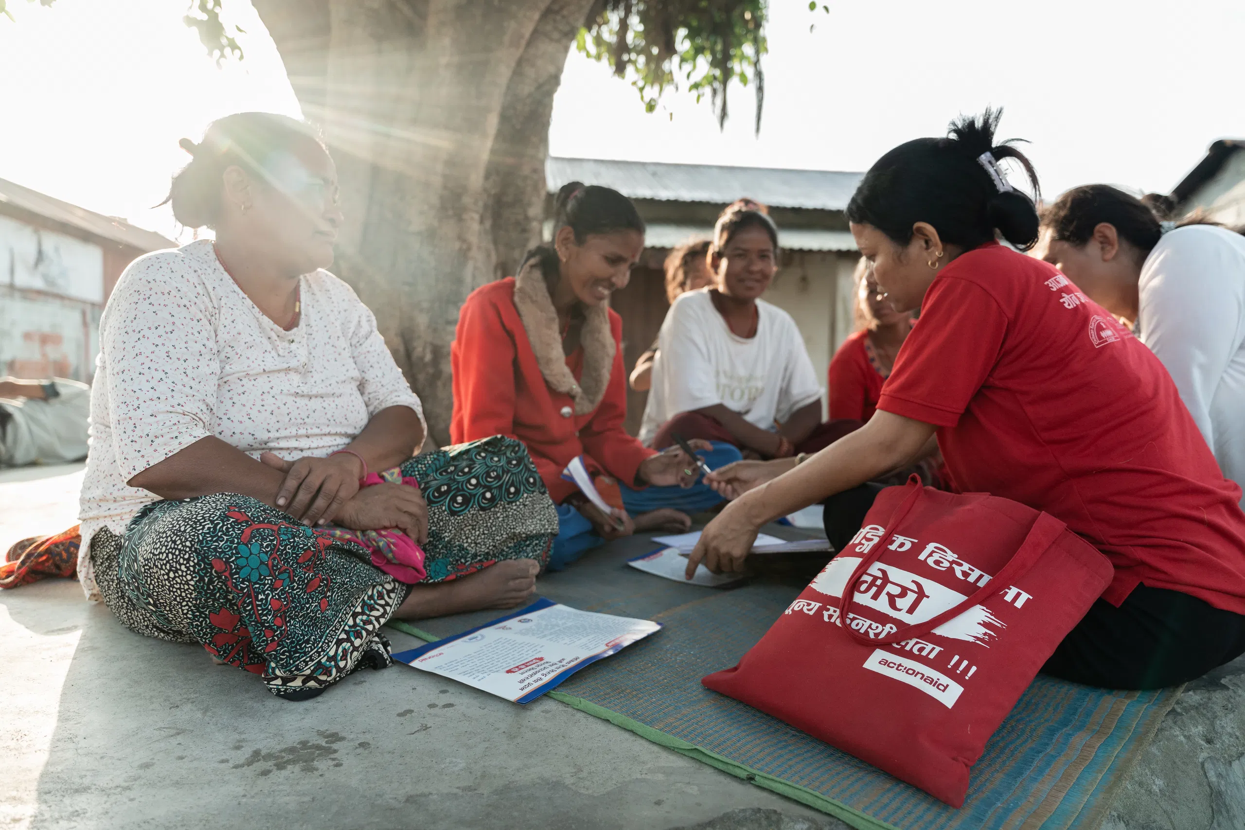 Women sitting together outdoors, engaging in a community meeting under a tree, sharing and discussing documents.