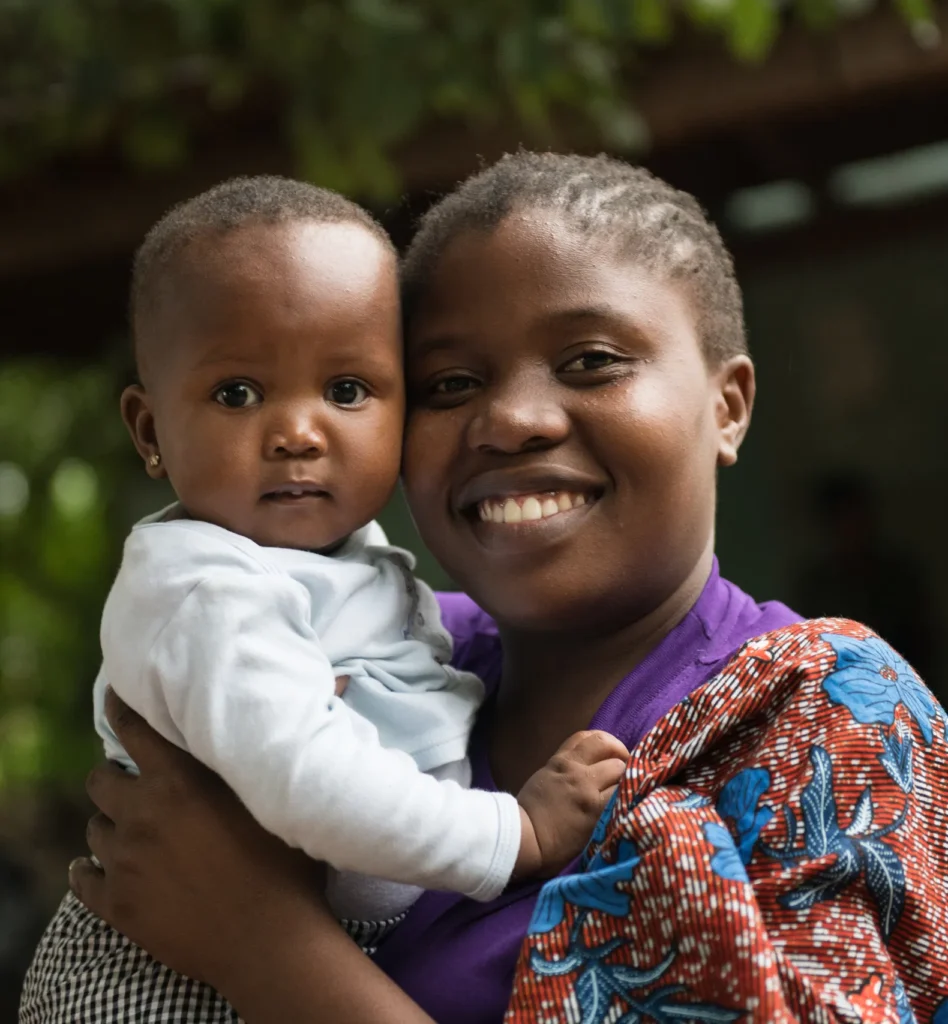 Smiling woman holds baby close in a warm embrace, outdoors with leafy background.