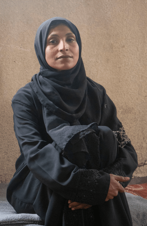 Smiling woman in black hijab and abaya sitting against a textured wall, showcasing cultural attire and elegance.
