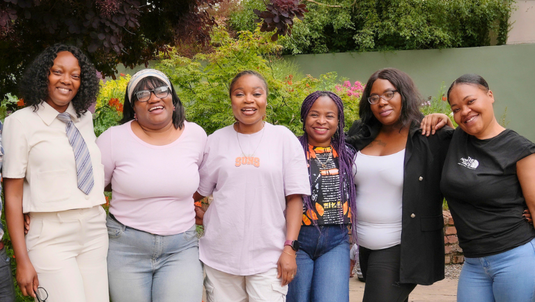 Group of six smiling women posing together outdoors in a garden setting.